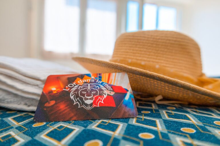 Carte d’Accueil « L’Or dans la Mer » posée sur un lit, avec chapeau de paille en arrière-plan.