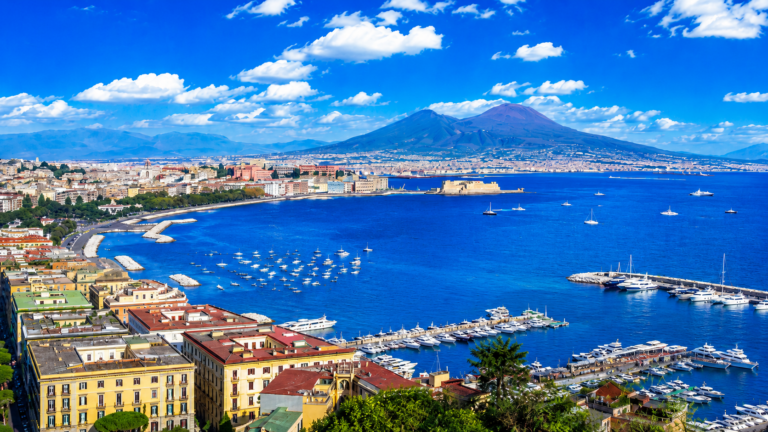 Vue panoramique sur le golfe de Naples, avec le Vésuve à l’arrière-plan, le front de mer, plusieurs ports de plaisance remplis de bateaux et une mer d’un bleu intense sous un ciel lumineux parsemé de nuages blanches.